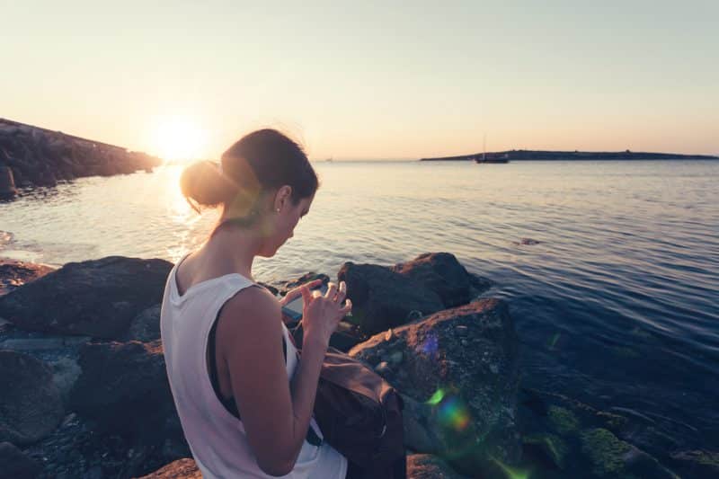 Young woman sitting at a beach at sunset using smartphone, Oshi app IBD management tool